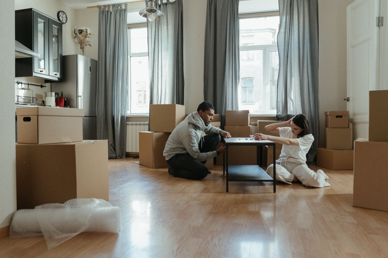 A young couple is putting together furniture in their new apartment, with cardboard boxes all around them.